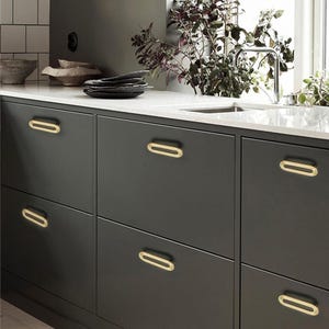 May include: Dark grey kitchen cabinets with oval gold-toned drawer pulls. The cabinets are set against a white countertop and a white tiled backsplash. A sink and tap are visible in the background.