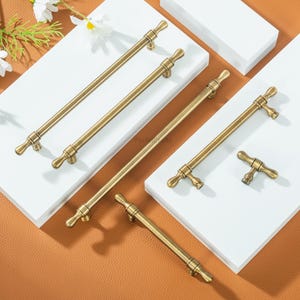 May include: A collection of antique brass cabinet pulls displayed on a white surface. The pulls feature a T-bar design with decorative ends. The hardware has a warm, golden hue, suggesting a vintage style. The pulls are of different lengths.