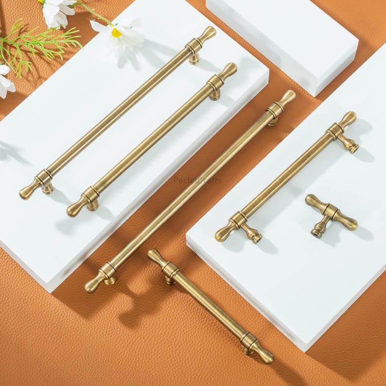 May include: A collection of antique brass cabinet pulls displayed on a white surface. The pulls feature a T-bar design with decorative ends. The hardware has a warm, golden hue, suggesting a vintage style. The pulls are of different lengths.