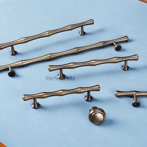 May include: A selection of antique brass cabinet hardware, featuring bamboo-style handles and a round knob. The hardware is displayed on a light blue surface, highlighting the detailed design and finish. The hardware is designed for cabinets and drawers.