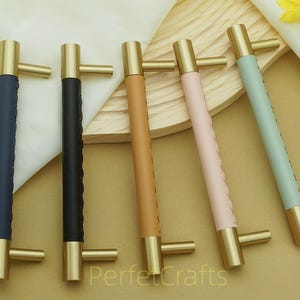 Puede incluir: Cinco tiradores de armario con herrajes de lat&oacute;n y mangos envueltos en cuero en azul marino, negro, marr&oacute;n, rosa y verde menta. Los tiradores est&aacute;n dispuestos sobre una superficie marr&oacute;n claro.