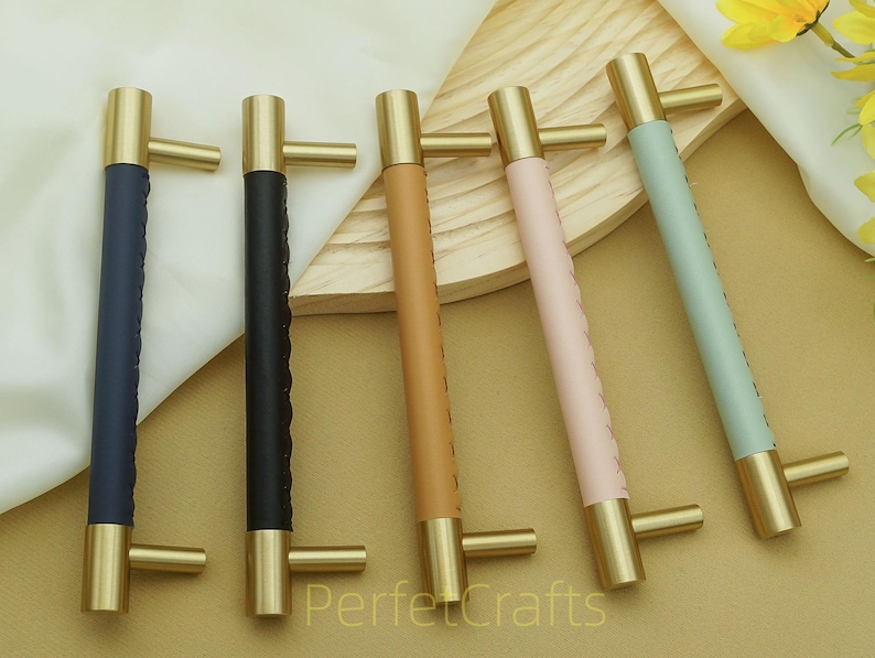 Puede incluir: Cinco tiradores de armario con herrajes de lat&oacute;n y mangos envueltos en cuero en azul marino, negro, marr&oacute;n, rosa y verde menta. Los tiradores est&aacute;n dispuestos sobre una superficie marr&oacute;n claro.