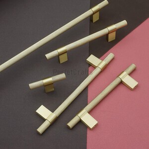 K&ouml;nnte beinhalten: Eine Sammlung goldfarbener Schrankgriffe mit einem strukturierten, rautenf&ouml;rmigen Griff. Die Griffe haben ein T-Stangen-Design und werden vor einem Hintergrund aus dunkelgrauen und rosa Oberfl&auml;chen pr&auml;sentiert. Die Hardware ist wahrscheinlich f&uuml;r M&ouml;bel oder Schr&auml;nke.