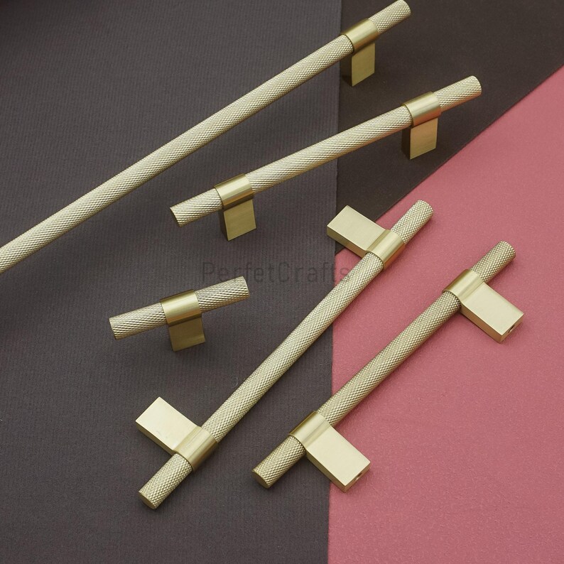 K&ouml;nnte beinhalten: Eine Sammlung goldfarbener Schrankgriffe mit einem strukturierten, rautenf&ouml;rmigen Griff. Die Griffe haben ein T-Stangen-Design und werden vor einem Hintergrund aus dunkelgrauen und rosa Oberfl&auml;chen pr&auml;sentiert. Die Hardware ist wahrscheinlich f&uuml;r M&ouml;bel oder Schr&auml;nke.