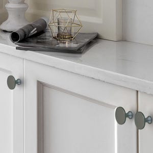 Op de afbeelding: Witte kastdeuren met grijze ronde knoppen. Een marmeren aanrechtblad bevat een opgerold tijdschrift, een gouden geometrische kandelaar met een glas en een decoratieve witte pilaar. De kastbeslag geeft de kamer een moderne uitstraling.