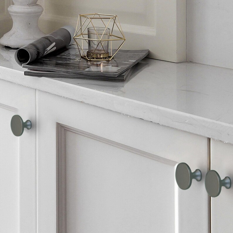 Op de afbeelding: Witte kastdeuren met grijze ronde knoppen. Een marmeren aanrechtblad bevat een opgerold tijdschrift, een gouden geometrische kandelaar met een glas en een decoratieve witte pilaar. De kastbeslag geeft de kamer een moderne uitstraling.