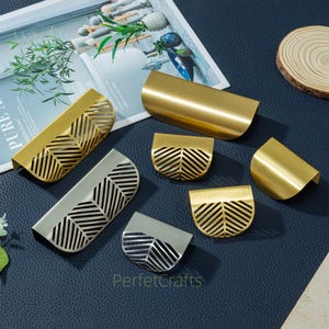 Op de afbeelding: Een verzameling gouden en zilveren metalen kastgrepen met een bladerenpatroon. De grepen zijn er in verschillende vormen en maten, waaronder rechthoekig en halfrond. De afbeelding staat op een donkerblauwe ondergrond.