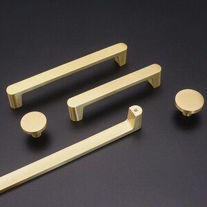May include: A selection of gold-coloured cabinet hardware, featuring two bar pulls, a long pull, and two round knobs. The hardware has a sleek, modern design with rounded edges, suitable for kitchen or bathroom cabinets.