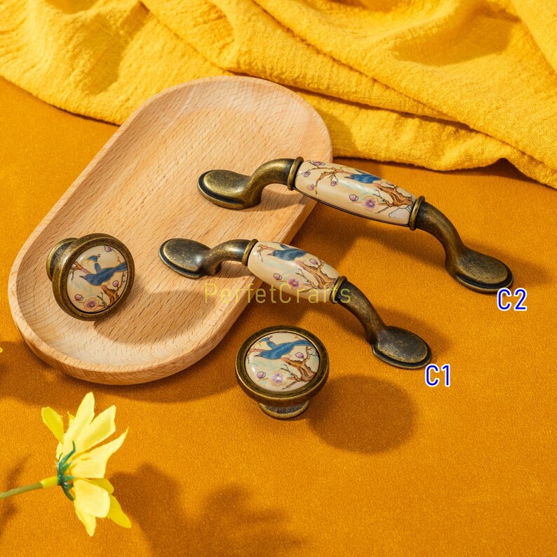 May include: A set of cabinet hardware featuring a bird and floral design. The set includes two handles and two knobs. The handles have a curved shape with a cream-colored ceramic center and bronze-colored metal ends. The knobs have a similar design.