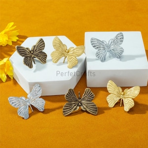 May include: Six decorative butterfly-shaped objects in various metallic finishes: bronze, gold, and silver. The butterflies are displayed on a white surface and a yellow-orange background. The text "PerfctCrafts" is visible.