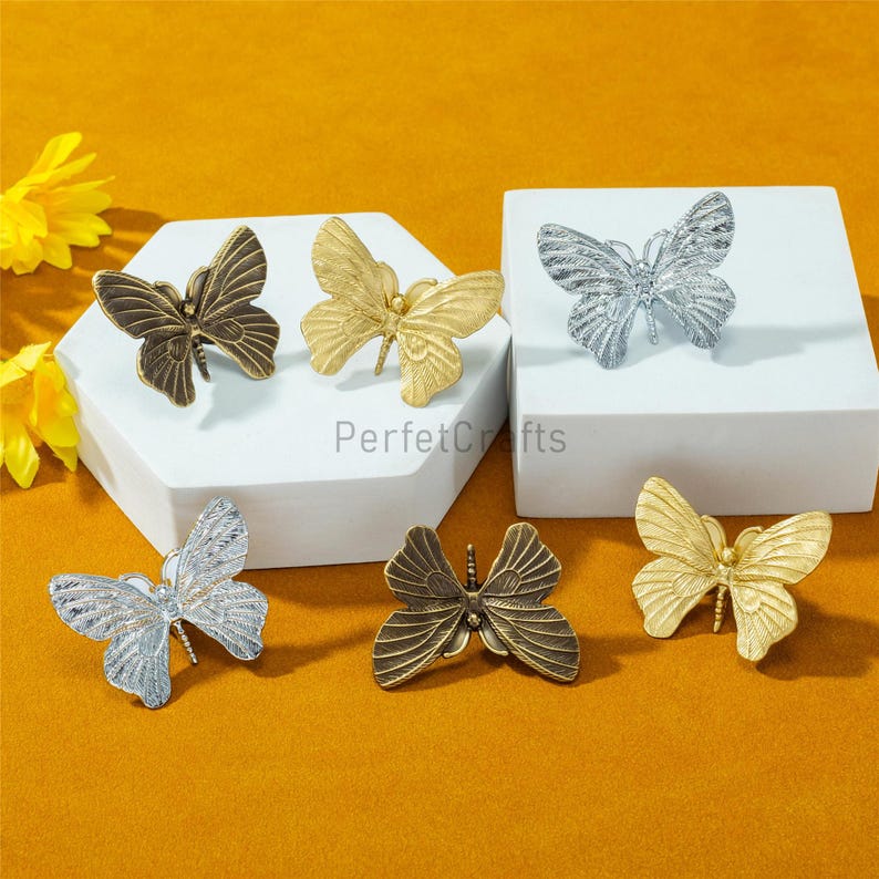May include: Six decorative butterfly-shaped objects in various metallic finishes: bronze, gold, and silver. The butterflies are displayed on a white surface and a yellow-orange background. The text "PerfctCrafts" is visible.