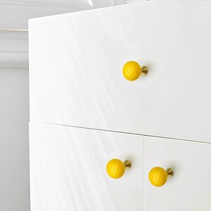 May include: White cabinet with three yellow round knobs. The knobs have a brass base and are evenly spaced on the cabinet doors. The cabinet has a glossy finish. The knobs are a cheerful accent.