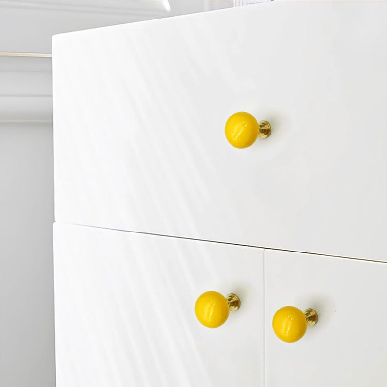 May include: White cabinet with three yellow round knobs. The knobs have a brass base and are evenly spaced on the cabinet doors. The cabinet has a glossy finish. The knobs are a cheerful accent.
