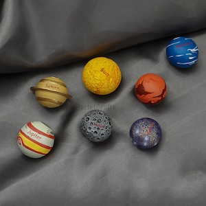May include: A collection of eight planet-themed spheres. The spheres are painted in various colors, including yellow, orange, blue, and gray, and labeled with the names of planets and the sun. The spheres are arranged on a gray surface.