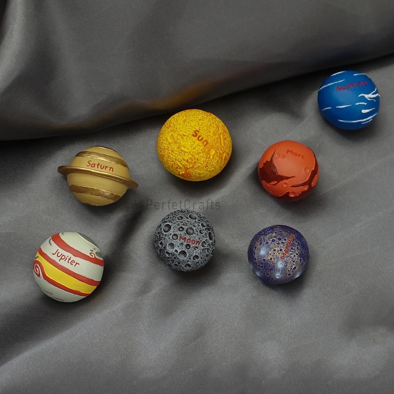 May include: A collection of eight planet-themed spheres. The spheres are painted in various colors, including yellow, orange, blue, and gray, and labeled with the names of planets and the sun. The spheres are arranged on a gray surface.
