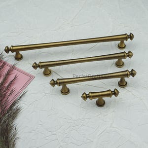 May include: Four antique brass cabinet pulls of varying sizes. Each pull features a cylindrical bar with decorative end pieces and a curved base. The pulls are arranged on a white textured surface. The text "PerfetcCrafts" is visible on the surface.