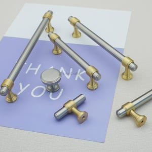 Peut inclure: Un ensemble de quincaillerie d'armoire, comprenant des poign&eacute;es argent&eacute;es et dor&eacute;es et un bouton. Les poign&eacute;es ont un design cylindrique avec des accents dor&eacute;s. Le bouton est rond avec une finition argent bross&eacute;. L'ensemble est sur fond violet et blanc.