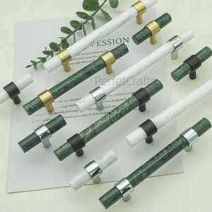 Op de afbeelding: Een verzameling kastgrepen met marmer en metalen accenten. De grepen hebben witte en groene marmeren cilinders met gouden, zilveren en zwarte metalen hardware. De afbeelding is op een witte achtergrond met tekst.