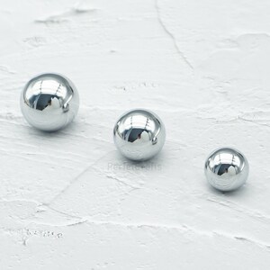 Op de afbeelding: Drie reflecterende, chroomkleurige bollen van verschillende grootte liggen op een witte, getextureerde ondergrond. De bollen hebben een gepolijste, spiegelachtige afwerking, die de omgeving weerspiegelt. De grootste bol is links, de kleinste rechts.