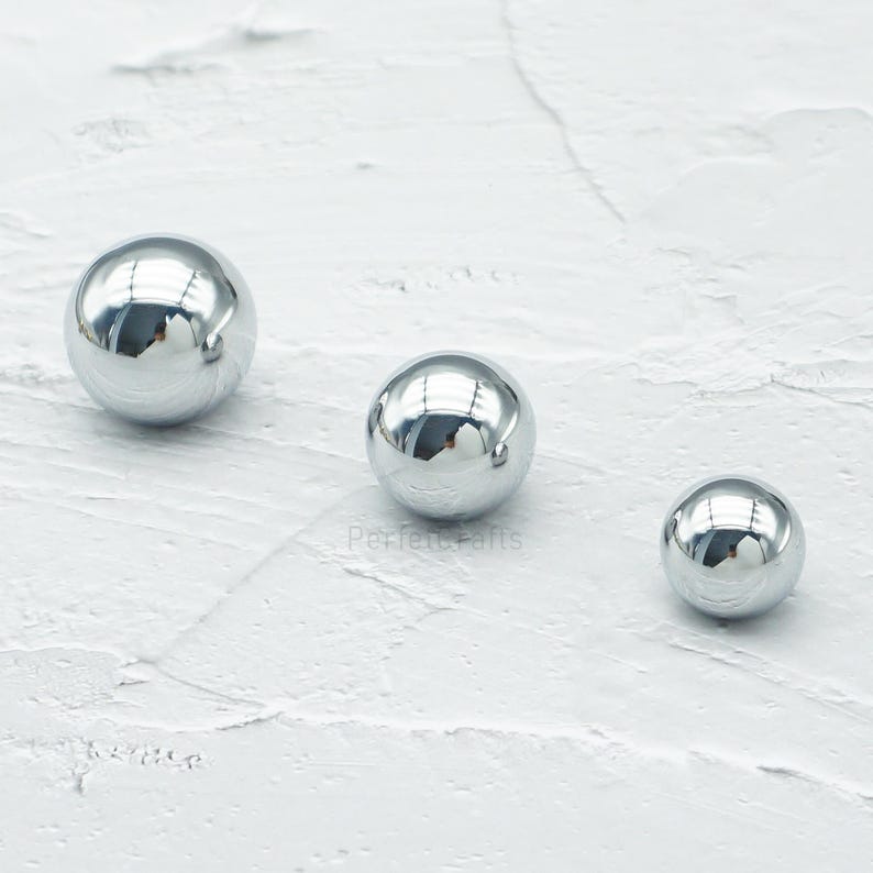 Op de afbeelding: Drie reflecterende, chroomkleurige bollen van verschillende grootte liggen op een witte, getextureerde ondergrond. De bollen hebben een gepolijste, spiegelachtige afwerking, die de omgeving weerspiegelt. De grootste bol is links, de kleinste rechts.