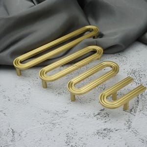 May include: Four gold cabinet pulls with an oval, grooved design. The pulls are arranged in a row, varying in size. The hardware has a matte finish and is set against a grey background.