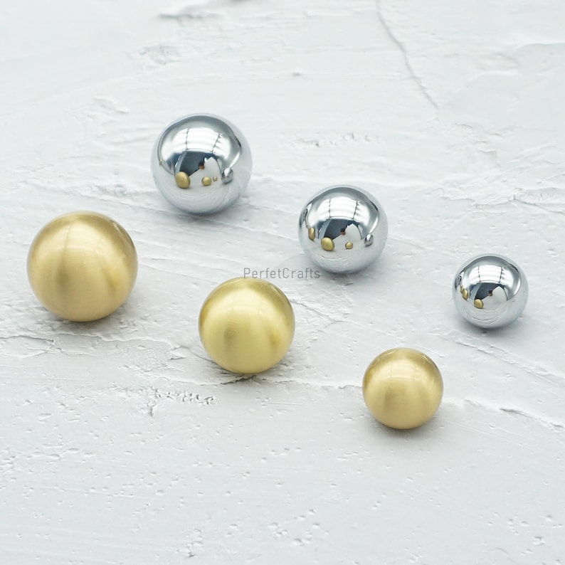 Op de afbeelding: Een assortiment metalen bollen in verschillende maten. Sommige bollen hebben een glanzende, zilveren afwerking, terwijl andere mat goudkleurig zijn. De bollen zijn gerangschikt op een getextureerd, lichtgekleurd oppervlak.