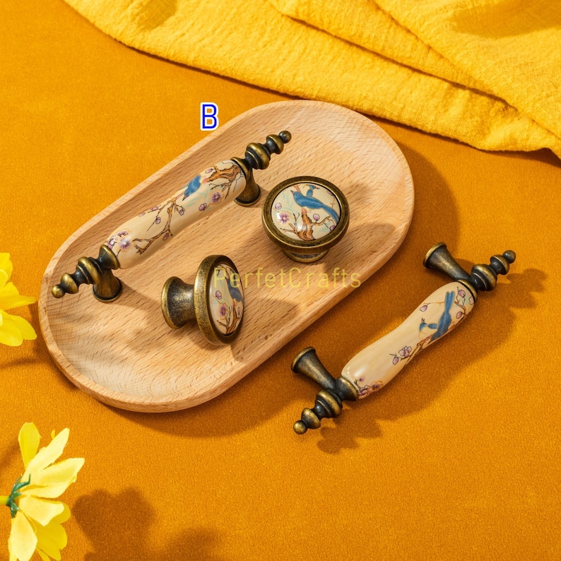 May include: A collection of antique-style cabinet hardware, including drawer pulls and knobs. The hardware features a cream-colored ceramic base with a blue bird and floral design, accented by bronze-colored metal. The items are arranged on a wooden tray.