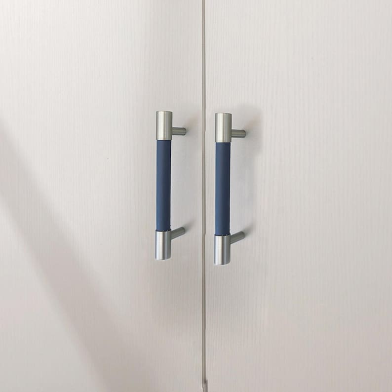 Puede incluir: Dos tiradores de armario con un dise&ntilde;o moderno. Cada tirador presenta un agarre cil&iacute;ndrico azul marino, flanqueado por extremos y postes de montaje de metal plateado cepillado. Montados en una puerta de armario blanca.