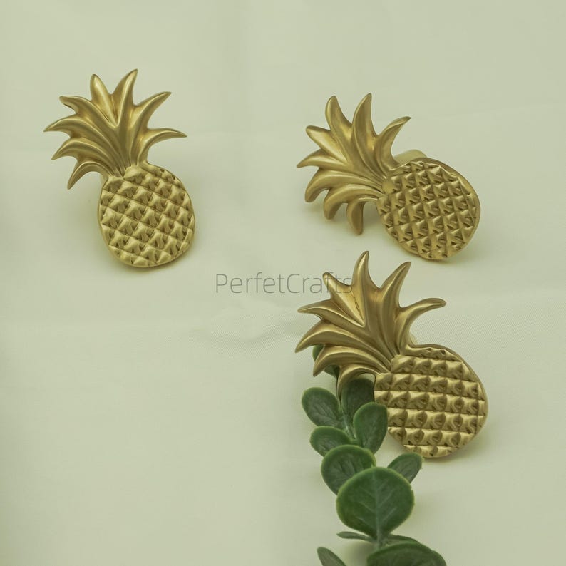 May include: Three gold-coloured pineapple-shaped cabinet knobs. The knobs have a textured surface and are arranged on a light background. The knobs are made of metal and are likely used for furniture or decorative purposes.