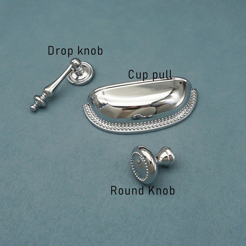 Może przedstawiać: Trzy błyszczące, srebrne elementy okuć do szafek: gałka kroplowa, uchwyt miseczkowy i okrągła gałka. Gałka kroplowa ma dekoracyjny trzon. Uchwyt miseczkowy ma koralikową krawędź. Okrągła gałka ma płaską, kropkowaną powierzchnię.