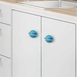 May include: Two blue planet-shaped cabinet knobs with white rings and the word "Uranus" printed on them. The knobs are attached to a white cabinet door. The cabinet is part of a kitchen setup with a light wood countertop and a stainless steel sink.