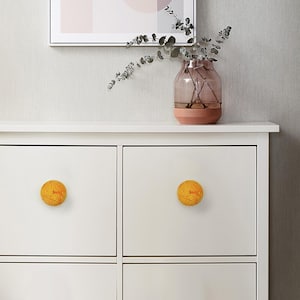May include: White cabinet with two yellow, spherical drawer pulls. Each pull has the word "Sun" in red. A vase with pink and brown tones sits on top of the cabinet, with a framed picture above.