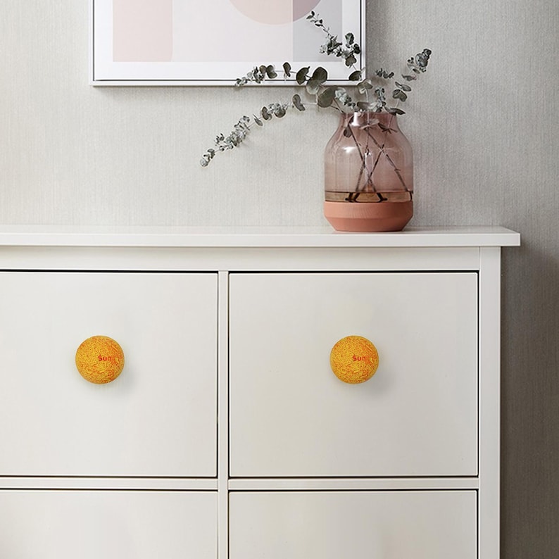 May include: White cabinet with two yellow, spherical drawer pulls. Each pull has the word "Sun" in red. A vase with pink and brown tones sits on top of the cabinet, with a framed picture above.