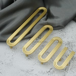 May include: Four gold cabinet pulls in different sizes, displayed on a textured surface. Each pull has a rounded, elongated shape with three parallel grooves. The pulls are made of metal and have a matte finish.