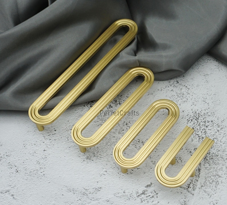 May include: Four gold cabinet pulls in different sizes, displayed on a textured surface. Each pull has a rounded, elongated shape with three parallel grooves. The pulls are made of metal and have a matte finish.