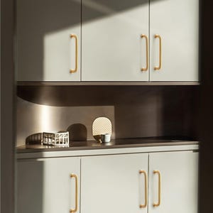 May include: A modern kitchen cabinet design featuring light grey cabinets with gold-coloured handles. The handles have a simple, curved design. The cabinets are arranged in a clean, minimalist style, with a shelf in between.