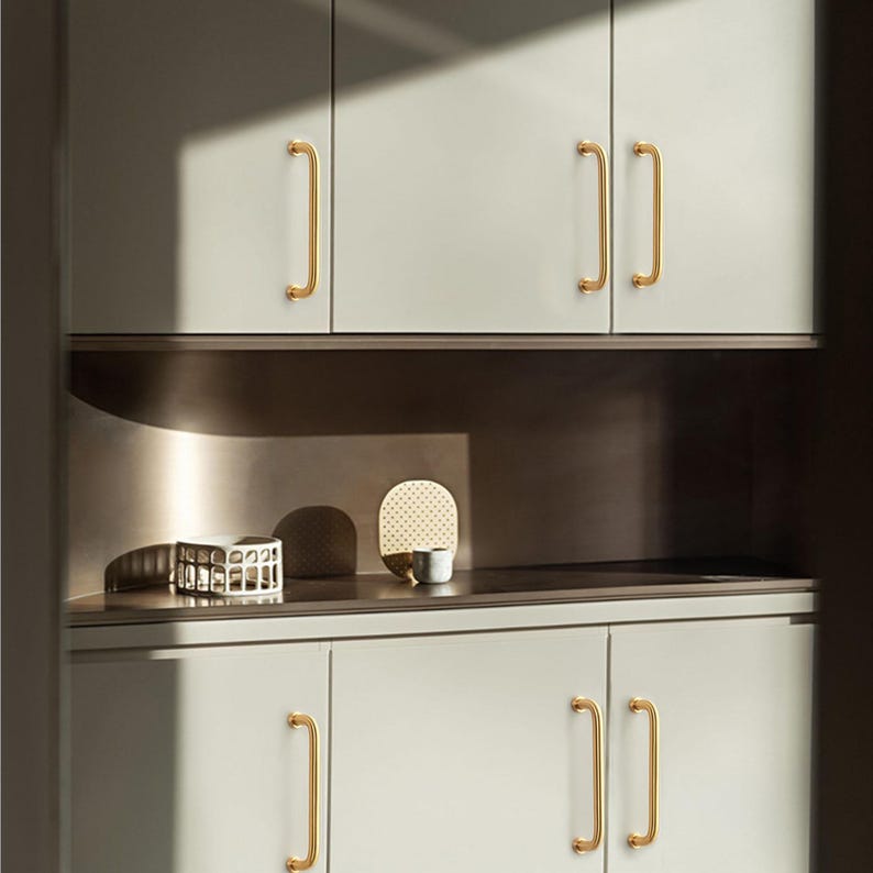 May include: A modern kitchen cabinet design featuring light grey cabinets with gold-coloured handles. The handles have a simple, curved design. The cabinets are arranged in a clean, minimalist style, with a shelf in between.