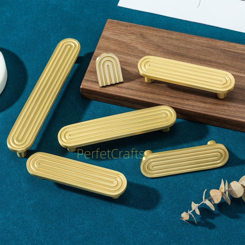 May include: Gold-colored cabinet hardware, including long pulls, small pulls, and a decorative arched piece. The hardware has a grooved design and is displayed on a blue surface and a wooden board. The text "PerfektCrafts" is visible.