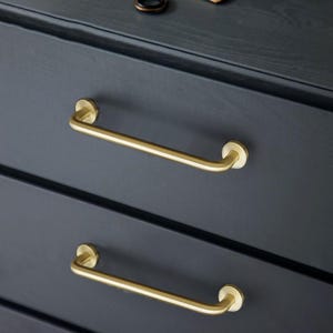 May include: Close-up of a dark grey chest of drawers featuring two brass bar handles. The handles are curved and attached to the drawer fronts with circular mounts. The wood grain is visible.