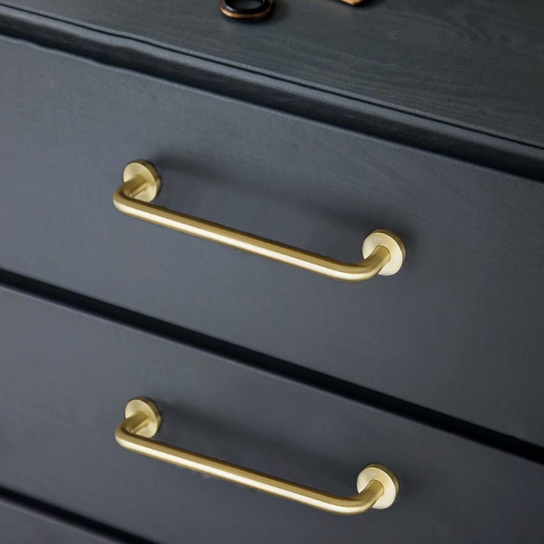 May include: Close-up of a dark grey chest of drawers featuring two brass bar handles. The handles are curved and attached to the drawer fronts with circular mounts. The wood grain is visible.