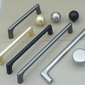 May include: An assortment of cabinet hardware in various metallic finishes. The selection includes several bar pulls in gold, black, and silver, along with matching knobs. The hardware is designed for furniture and cabinet doors.