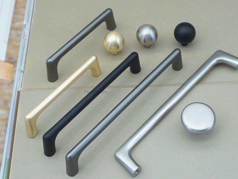 May include: An assortment of cabinet hardware in various metallic finishes. The selection includes several bar pulls in gold, black, and silver, along with matching knobs. The hardware is designed for furniture and cabinet doors.
