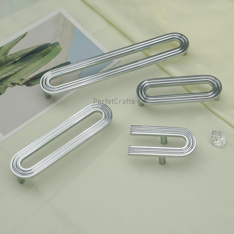 May include: A selection of silver cabinet pulls in different sizes. The pulls feature a grooved, oval design with a curved handle. The image includes the text "PerfetcCrafts". The hardware is displayed on a light green surface.