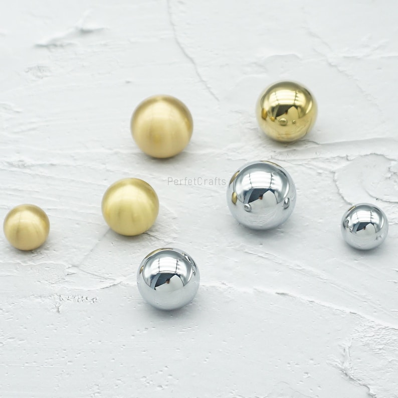 Op de afbeelding: Een assortiment bolvormige kastknoppen in gouden en zilveren afwerkingen. De knoppen vari&euml;ren in grootte, van klein tot middelgroot. De metalen knoppen zijn gepresenteerd op een getextureerd, gebroken wit oppervlak.