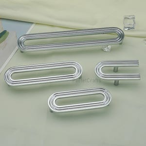 May include: A selection of silver cabinet pulls and handles in different sizes and shapes. The hardware has a grooved, oval design with a modern look. The image shows a long pull, two oval pulls, and a U-shaped handle. These are suitable for various furniture.