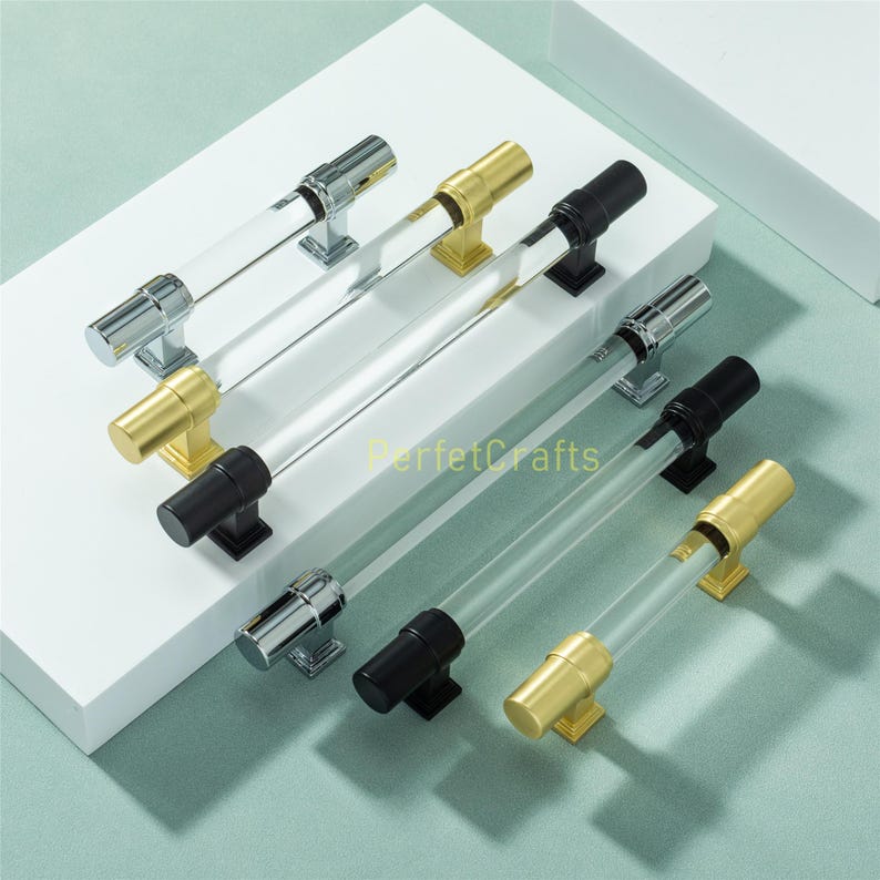 May include: A collection of cabinet pulls with clear acrylic rods and metal end caps. The metal finishes include chrome, gold, and black. The pulls are arranged on a white surface, showcasing their design and variety.