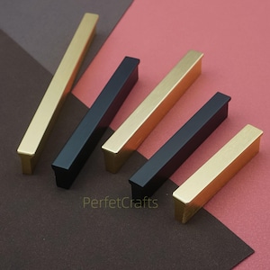 May include: An assortment of rectangular cabinet pulls in gold and black. The pulls are arranged diagonally across a two-tone brown and red surface. The pulls have a simple, modern design, with a rectangular bar and a small, square base.