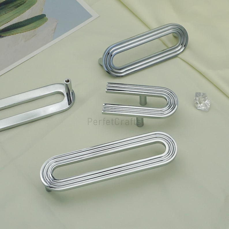 May include: Several polished silver cabinet pulls are displayed, featuring oval and U-shaped designs with a ribbed texture. The image also includes a small, clear crystal and the text "PerfecCrafts".