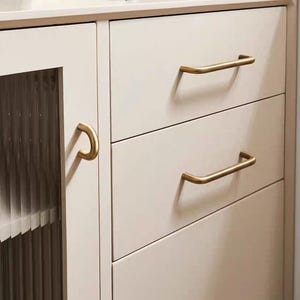 K&ouml;nnte beinhalten: Nahaufnahme eines cremefarbenen Schranks mit drei Schubladen und einer Glast&uuml;r. Die Schubladen haben goldfarbene, gebogene Stangengriffe. Der Schrank hat eine neutrale Farbe und eignet sich f&uuml;r verschiedene Einrichtungsstile.