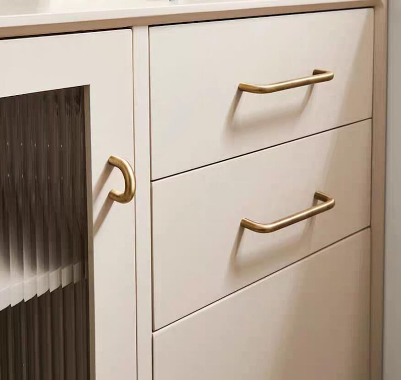 K&ouml;nnte beinhalten: Nahaufnahme eines cremefarbenen Schranks mit drei Schubladen und einer Glast&uuml;r. Die Schubladen haben goldfarbene, gebogene Stangengriffe. Der Schrank hat eine neutrale Farbe und eignet sich f&uuml;r verschiedene Einrichtungsstile.